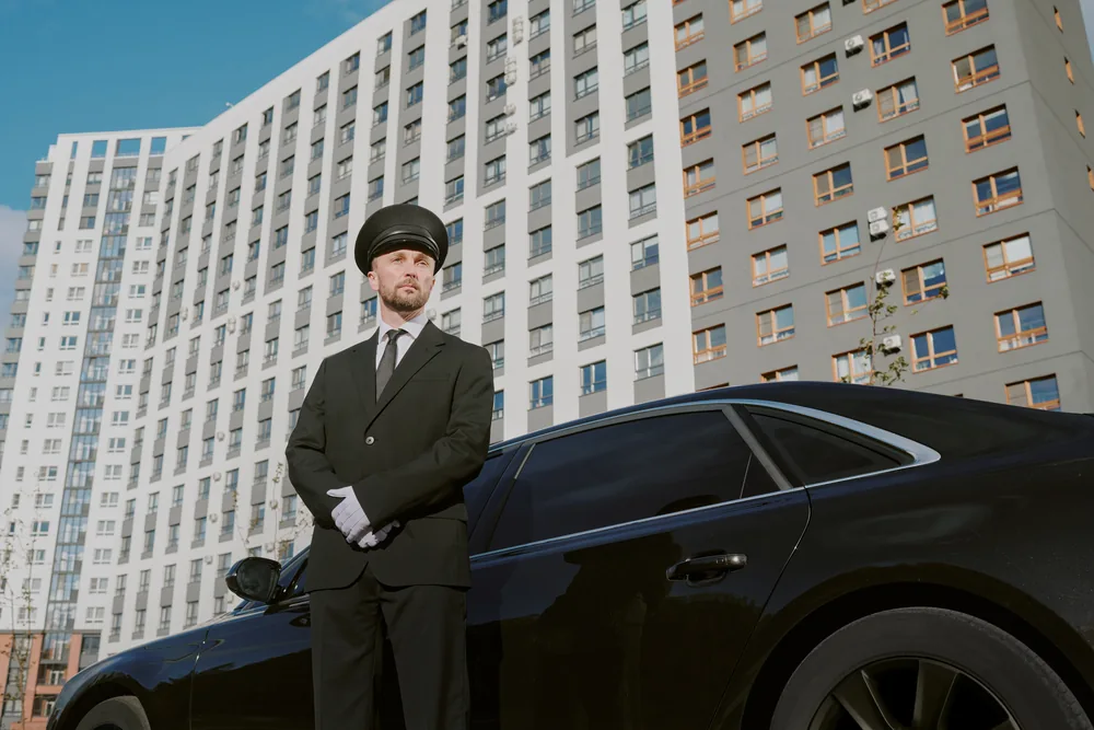 A uniformed chauffeur standing beside a black luxury sedan